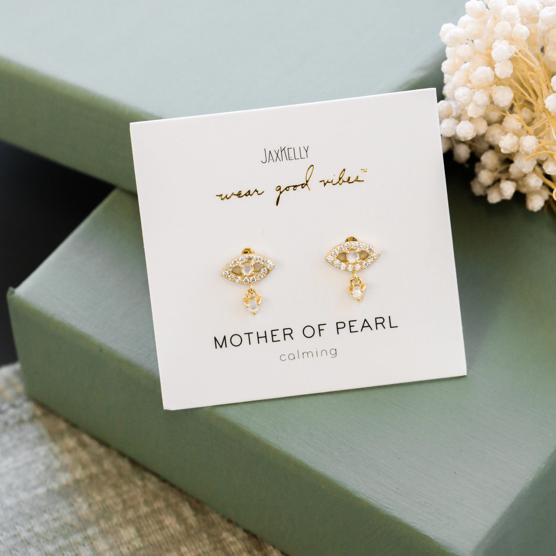 Mother of Pearl Evil Eye Drop earrings with gold, pearl, and CZ accents displayed on a white JaxKelly card labeled “calming, wear good vibes,” set atop green gift boxes with white dried flowers nearby.