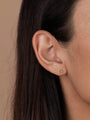 Close-up of a person's ear with straight brown hair, wearing the Cross Stud—an 18k gold plated earring on sterling silver posts. The background is plain beige.