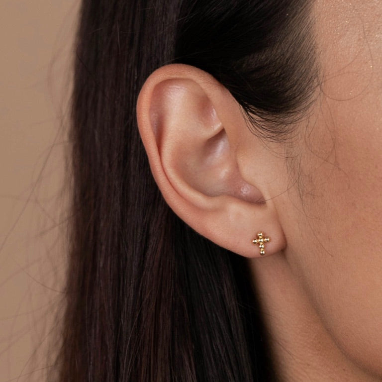 Close-up of a person's ear with straight brown hair, wearing the Cross Stud—an 18k gold plated earring on sterling silver posts. The background is plain beige.