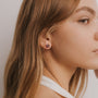 A close-up side profile of a young woman with light brown hair in a white top, wearing the Amethyst Gemstone Prong - Healing earring. The flower-shaped stud features an amethyst stone, set against a plain, light-colored background.
