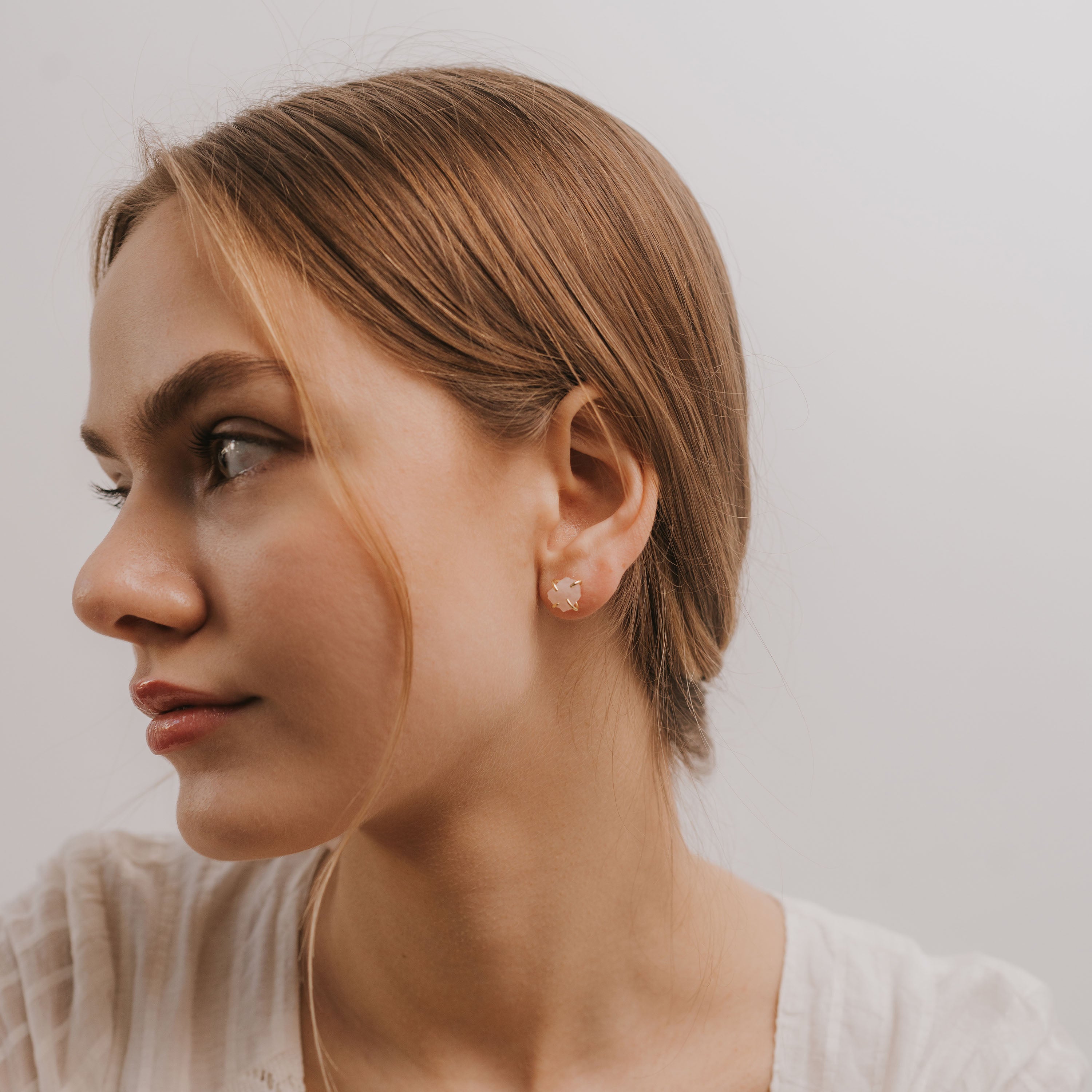 A young woman with light brown hair tied back looks left, wearing a white top and the Rose Quartz Gemstone Prong - Love earrings. The small, round rose quartz stones are set in sterling silver. The background is plain and light-colored.