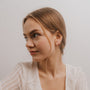 A young woman with light brown hair, styled in loose strands, gazes to the side. She wears a white semi-sheer top and the Rose Quartz Gemstone Prong - Love sterling silver stud earrings against a plain light background.