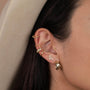 A close-up of a person’s ear wearing multiple gold-plated earrings, including the Pavé Snake stud with sparkling CZ stones, hoops, and a cuff. They have dark hair tucked behind their ear and are wearing a light-colored hat.