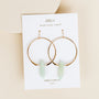 The Amazonite Hoop - Hope features gold plated hoops with light green amazonite crystal pendants, displayed on a white JaxKelly Wear Good Vibes™ card against a beige background.