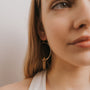A close-up of a young woman with long blonde hair, wearing a white top and JaxKelly’s hypoallergenic Citrine Hoop - Success earrings. The softly lit plain background highlights the elegance of these gold hoops with clear crystal pendants.