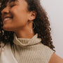 A woman with curly hair smiles with her eyes closed, wearing a sleeveless beige turtleneck and the Clear Quartz Hoop - Healing earring, featuring a handcrafted crystal and vertical bar detail. The background is plain and light-colored.