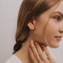 A young woman with light brown hair in a loose braid wears the Rose Quartz Hoop - Love earrings, gently touching one as she faces sideways against a neutral background, highlighting the unique beauty of her handcrafted jewelry.