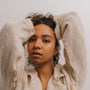 A woman with curly dark hair, dressed in a textured cream sweater and wearing Smoky Quartz Hoop - Protection earrings, poses with her arms raised and hands in her hair against a plain light background.