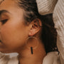 A close-up of a curly-haired person wearing the Smoky Quartz Hoop – Protection: a large gold hoop earring with a smoky quartz pendant. They wear a cream textured top, arm raised near their head, highlighting this handcrafted jewelry.