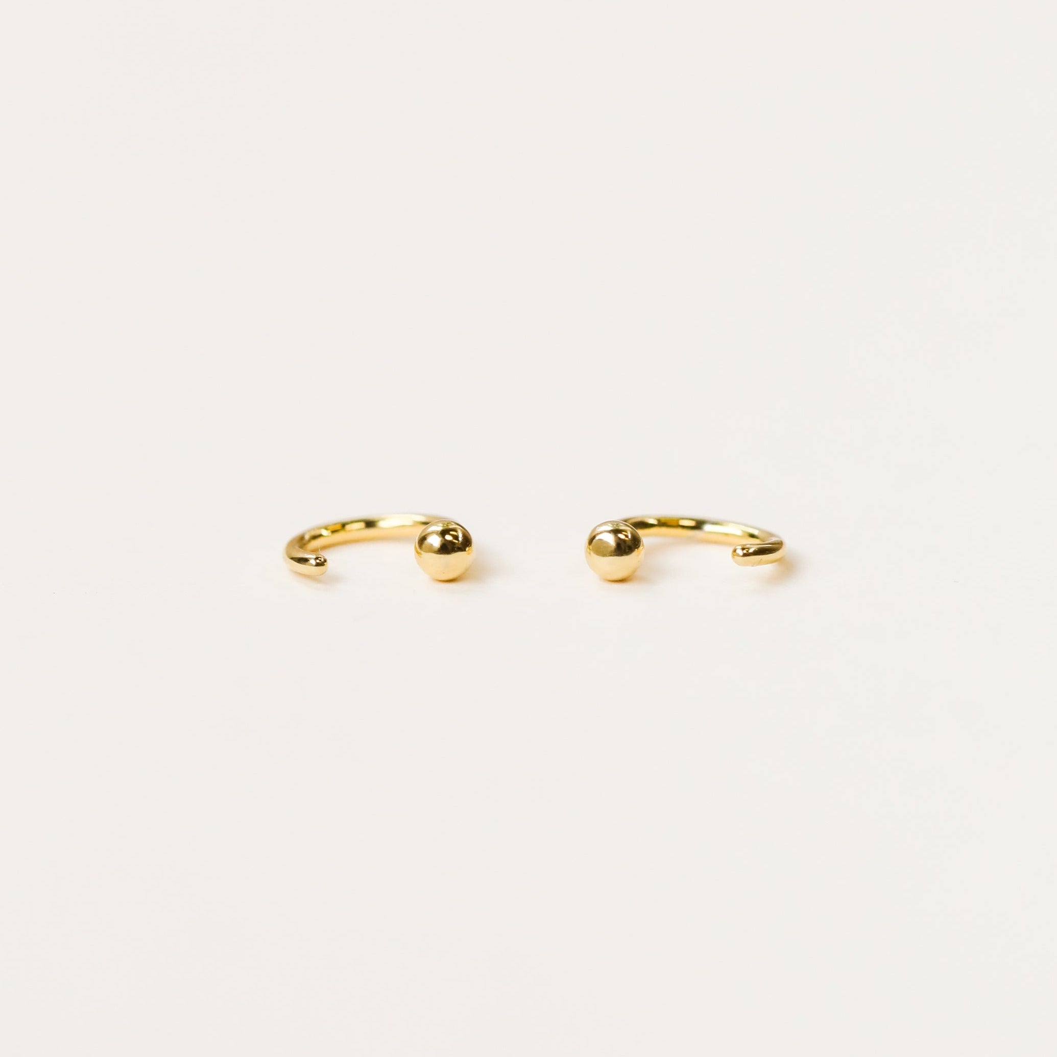 The Gold Sphere Huggie is a pair of small, open gold hoops each featuring a single round bead at one end, shown on a plain white background.