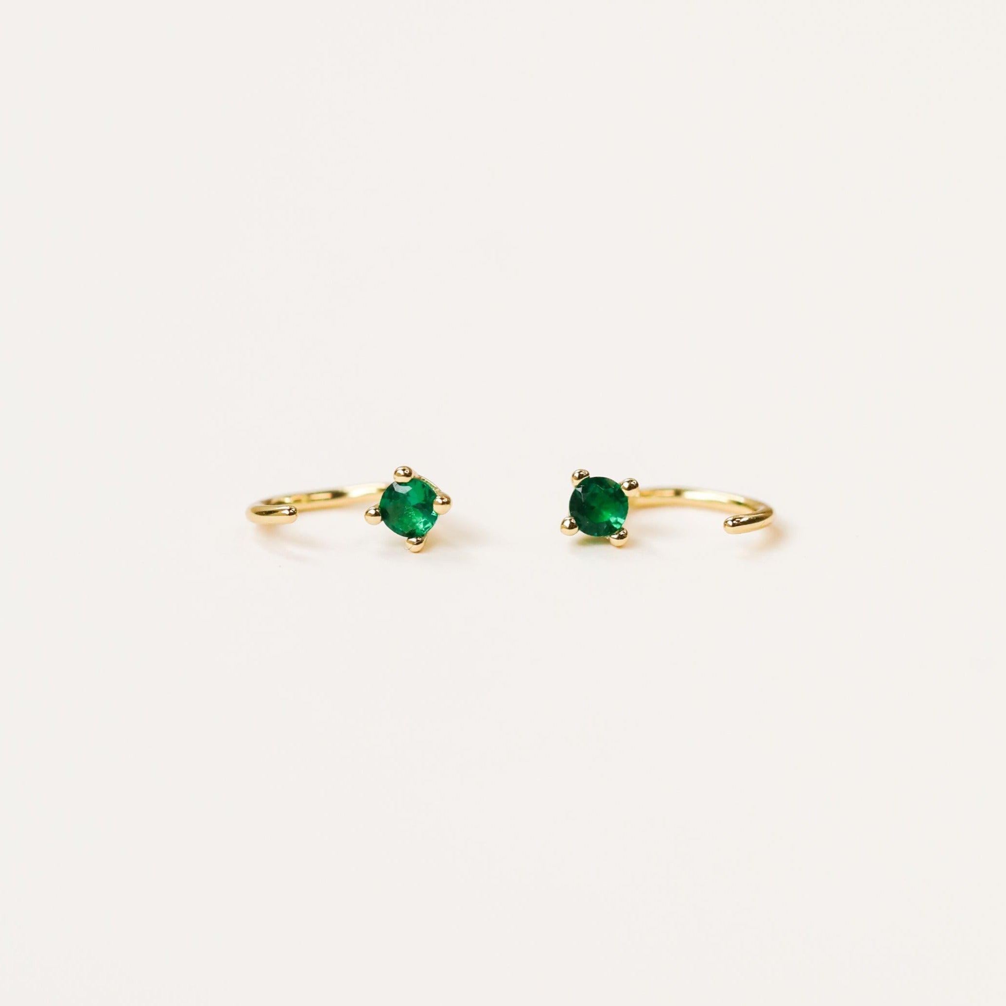 The Emerald CZ Huggie features delicate 18k gold plated huggie hoops, each set with a small, round emerald CZ stone in a four-prong setting, shown against a plain white background.