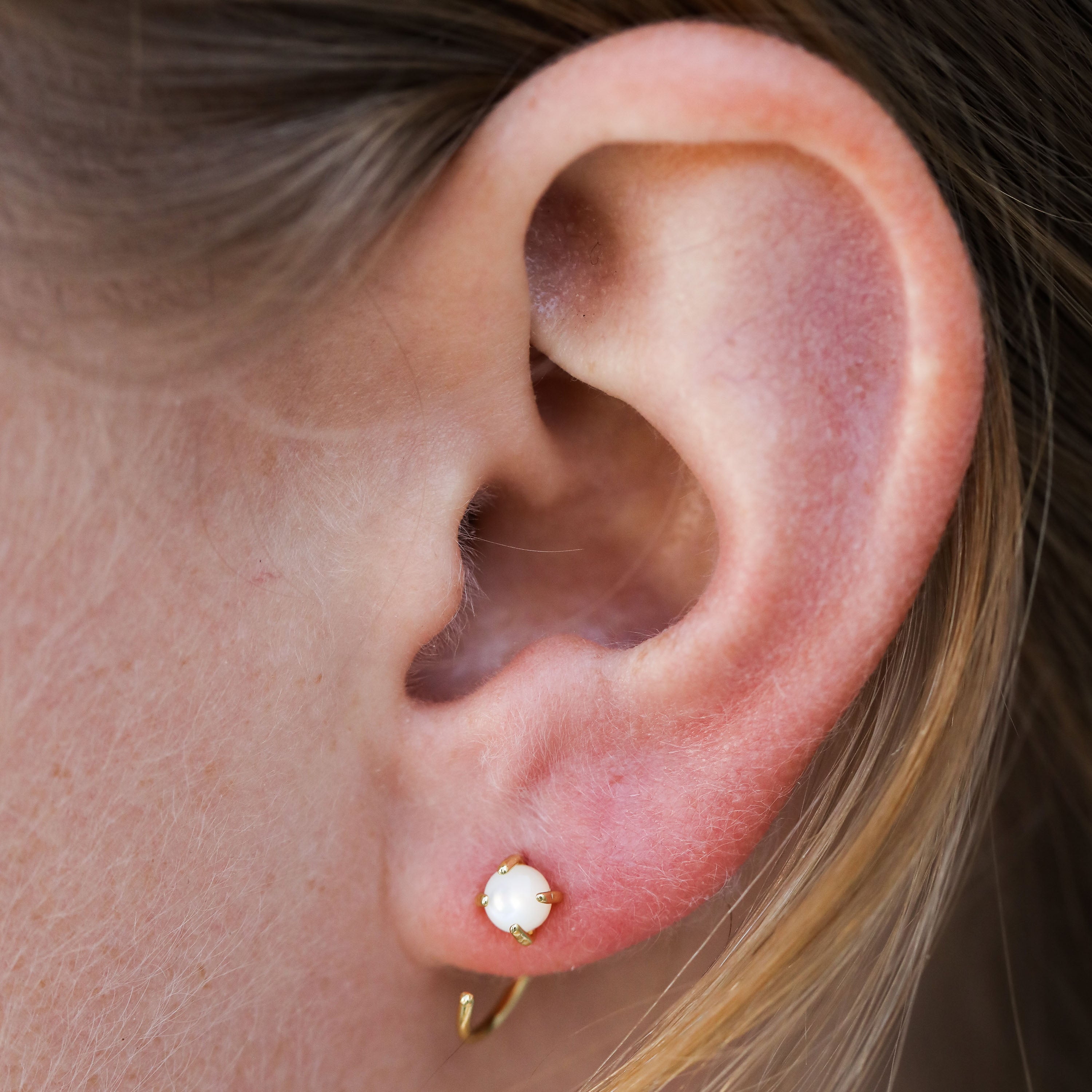 Close-up of a person’s ear wearing the Mother of Pearl Huggie—a small gold huggie earring with a white Mother of Pearl stone. Light brown hair is partially visible.