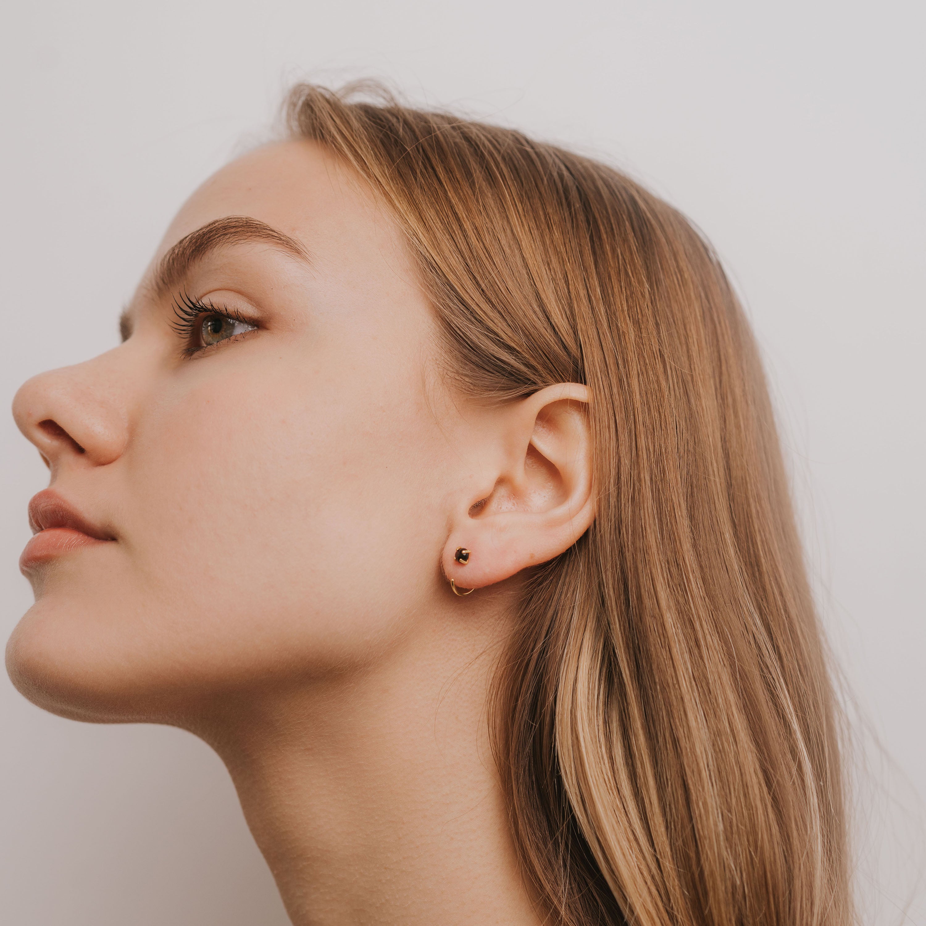 A young woman with light brown hair gazes upward in side profile, wearing the Obsidian Huggie—an 18k gold-plated hoop earring—in her left ear, set against a plain white background.