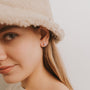 A woman with long blonde hair wears a beige fuzzy bucket hat and Fire Opal Huggie earrings. She smiles slightly at the camera against a plain light background.