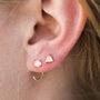 Close-up of an ear adorned with dainty earrings: a round stud, a triangular stud, and the Rose Quartz Huggie in the lower lobe. The person's light brown hair frames the edge.