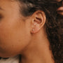 Close-up of a left ear wearing the Rose Quartz Huggie, a delicate gold earring. The person’s curly brown hair is pulled back, showing smooth skin and a neutral background—ideal for those who love dainty jewelry.