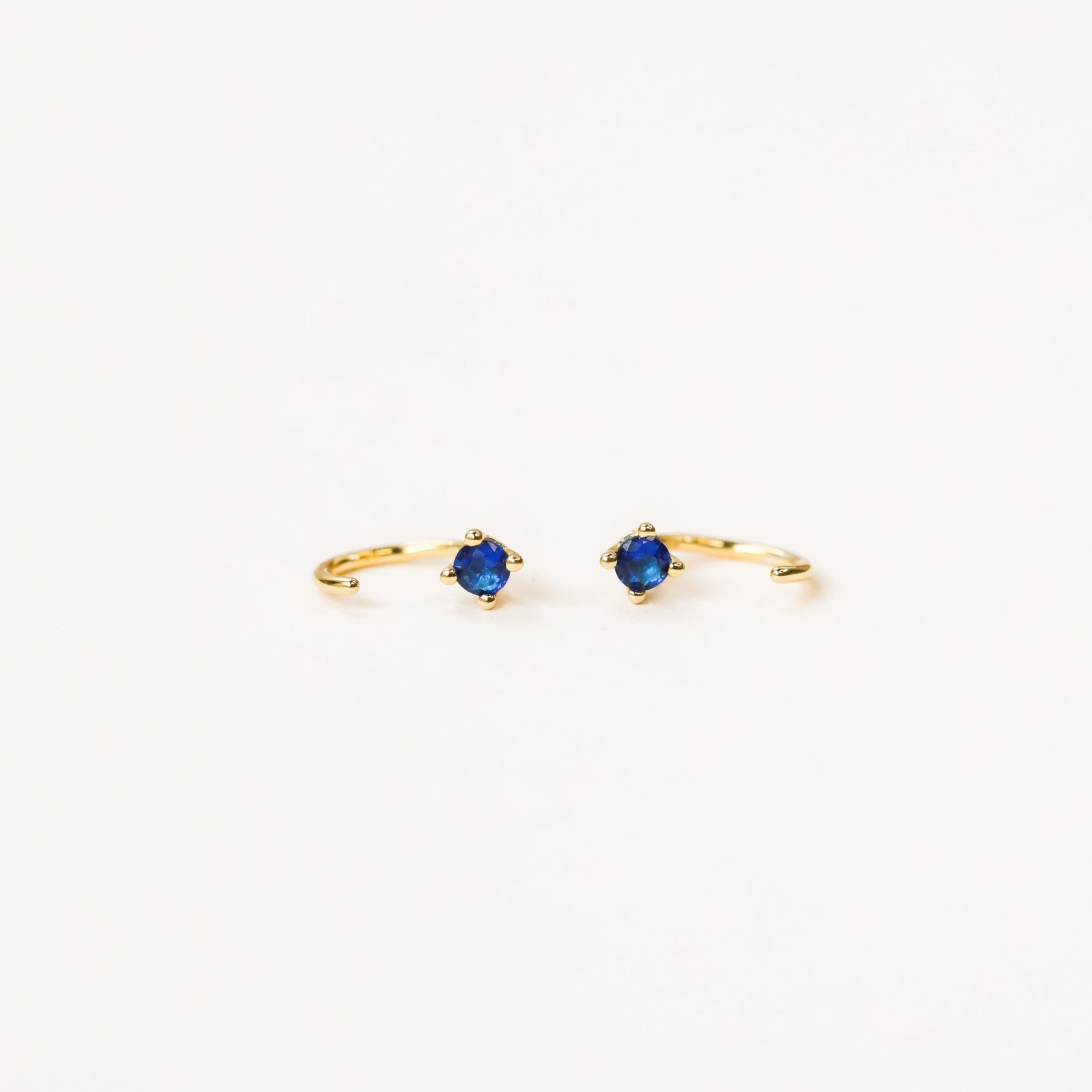 The Sapphire CZ Huggie features delicate gold open hoops with a single round blue cz stone at the center, shown on a plain white background.
