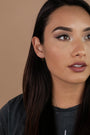 A woman with long dark hair and winged eyeliner looks off to the side. She wears a dark shirt, a pair of Stud Chain Huggie earrings, and stands against a neutral background.