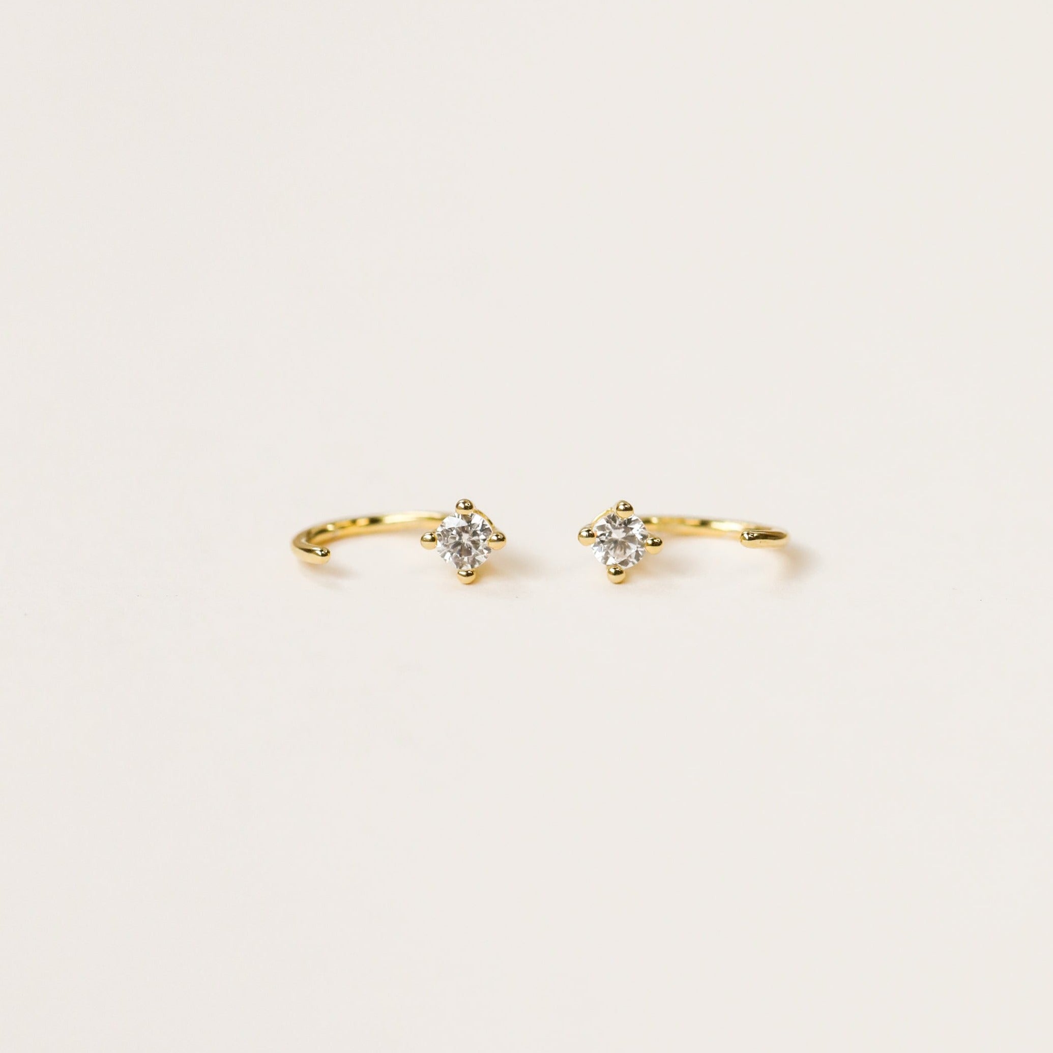 The White CZ Huggie is a pair of dainty gold-plated earrings, each featuring a small, round, clear gemstone at the center, displayed on a plain light background.