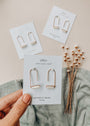 A hand holds a card showcasing Mother of Pearl Gemstone Drop - Calming earrings. Two similar sets are displayed on cards on a beige surface, with a green cloth and dried white flowers artfully arranged nearby.