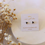 A pair of Black Tourmaline Mini Energy Gems - Strength studs with sterling silver posts on a white card with gold lettering, shown beside small white and yellow dried flowers against a light, neutral background.