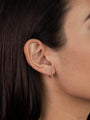 Close-up of a woman's ear with straight brown hair, wearing Tiny Ball Hoop gold-plated earrings with sterling silver posts, highlighting their elegant design and the side of her face.
