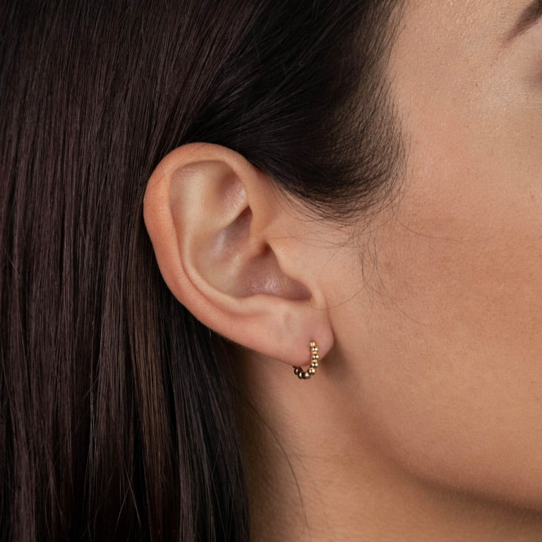 Close-up of a woman's ear with straight brown hair, wearing Tiny Ball Hoop gold-plated earrings with sterling silver posts, highlighting their elegant design and the side of her face.