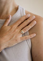 A person wearing a ribbed, sleeveless top rests their hand on their chest, showcasing the Opal Crown Ring—an 18k gold plated piece with a floral design and opal gemstone—on their ring finger.