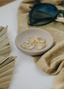 A small beige dish holds four gold rings, including the Mother of Pearl Signet Ring, displayed on tan fabric beside folded sunglasses and dried palm leaves.