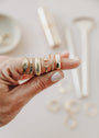 A hand with pale nail polish models five unique gold rings, including the Ridged Ring—a stackable band with 18k gold plating—while blurred gold rings and jewelry tools appear softly in the background.