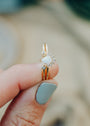 A hand with light blue nails holds the Opal Burst Ring—an 18k gold plated piece featuring a lab-grown Opal center, two small diamonds, and a cluster of accent stones.