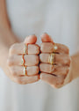 Two fists display several gold-plated rings, including the Half Pavé Ring adorned with sparkling stones and unique designs, all set against a soft, blurred white background.