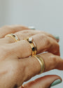 A close-up of a hand with light green nail polish showcases several gold rings, including the Ridged Ring and one with a small white stone—all featuring 18k gold plating—against a light, neutral background.
