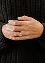 A person in a black textured sweater showcases their hands with two gold rings: the Mother of Pearl Signet Ring featuring 18k gold plating and a round stone, and another with a beaded design. Their nails are short and natural.