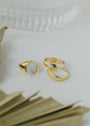 Three gold rings—a stackable 18k gold plated band with a white stone, a small white oval ring, and the Ridged Ring—are displayed on a white surface with blurred glass and dried palm leaf in the background.