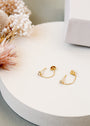 The Stud Chain Huggie, a pair of delicate 18k gold plated chain earrings with small gemstones, is displayed on a round white surface beside soft pink and white dried flowers and a white box.