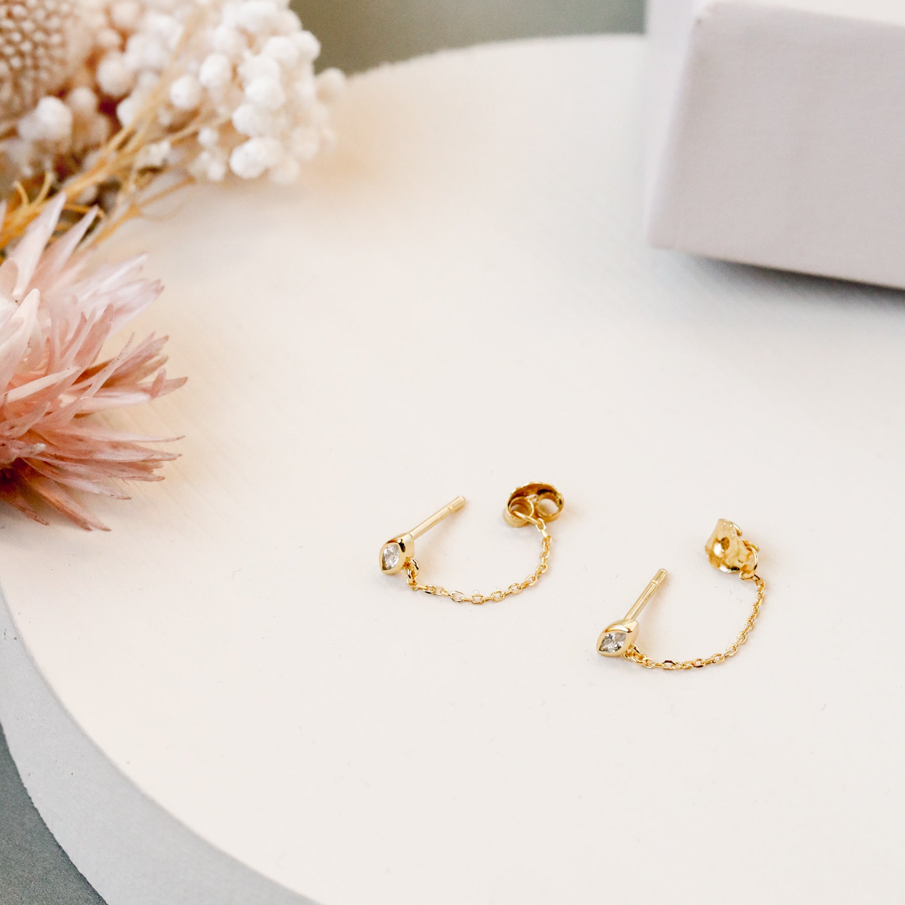 The Stud Chain Huggie, a pair of delicate 18k gold plated chain earrings with small gemstones, is displayed on a round white surface beside soft pink and white dried flowers and a white box.