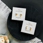 Two pairs of gold-plated sterling silver earrings with amazonite rest on white cards labeled "AMAZONITE hope" atop a black plate and grey fringed fabric. One pair is lips-shaped, the other is the Amazonite Moon Drop design.