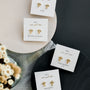 The Mother of Pearl Evil Eye Drop gold earrings with gemstone accents are displayed on a white card labeled "Mother of Pearl," arranged on a circular surface next to a vase of white flowers.