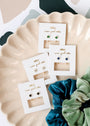 Four pairs of Sapphire CZ Huggie earrings with 18k gold plating are displayed on white cards with gold writing, set on a cream scalloped dish beside teal and green velvet scrunchies on a patterned surface.