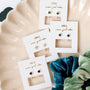 Four pairs of White CZ Huggie earrings on white cards with gold writing, arranged on a cream plate with green and blue velvet nearby. Each features unique colored stones and delicate gold-plated designs.