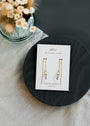 JaxKelly’s Gemstone Threader - Clear Quartz earrings are shown on a branded card that reads "Wear Good Vibes, Clear Quartz, cleansing," resting on a black plate next to a vase of dried flowers.