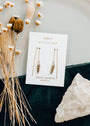 JaxKelly’s Gemstone Threader - Smoky Quartz earrings, featuring 18k gold plating and inscribed with “Wear Good Vibes™” and “Protection,” are shown on a branded card, beside dried flowers and a large quartz crystal on velvet.