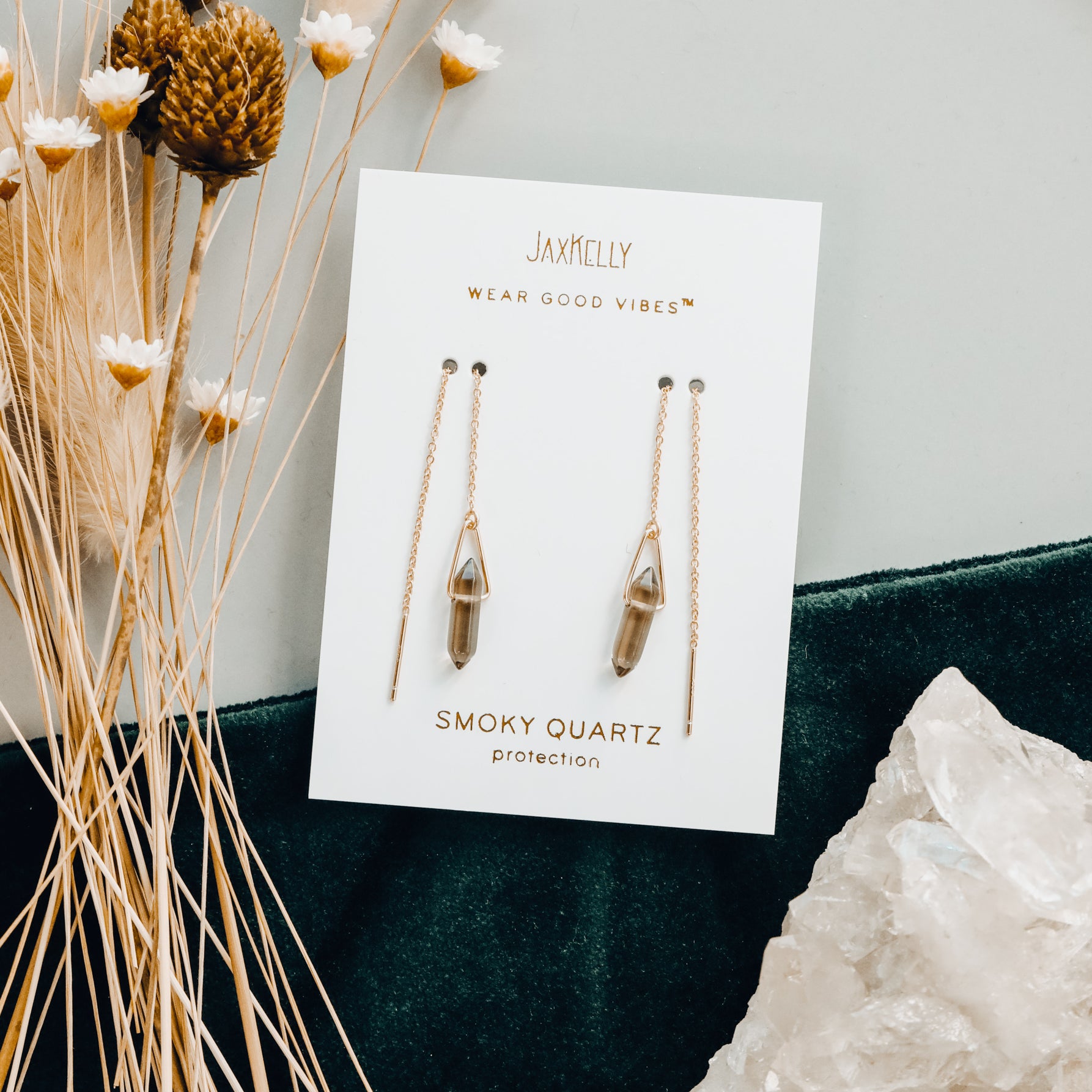 JaxKelly’s Gemstone Threader - Smoky Quartz earrings, featuring 18k gold plating and inscribed with “Wear Good Vibes™” and “Protection,” are shown on a branded card, beside dried flowers and a large quartz crystal on velvet.