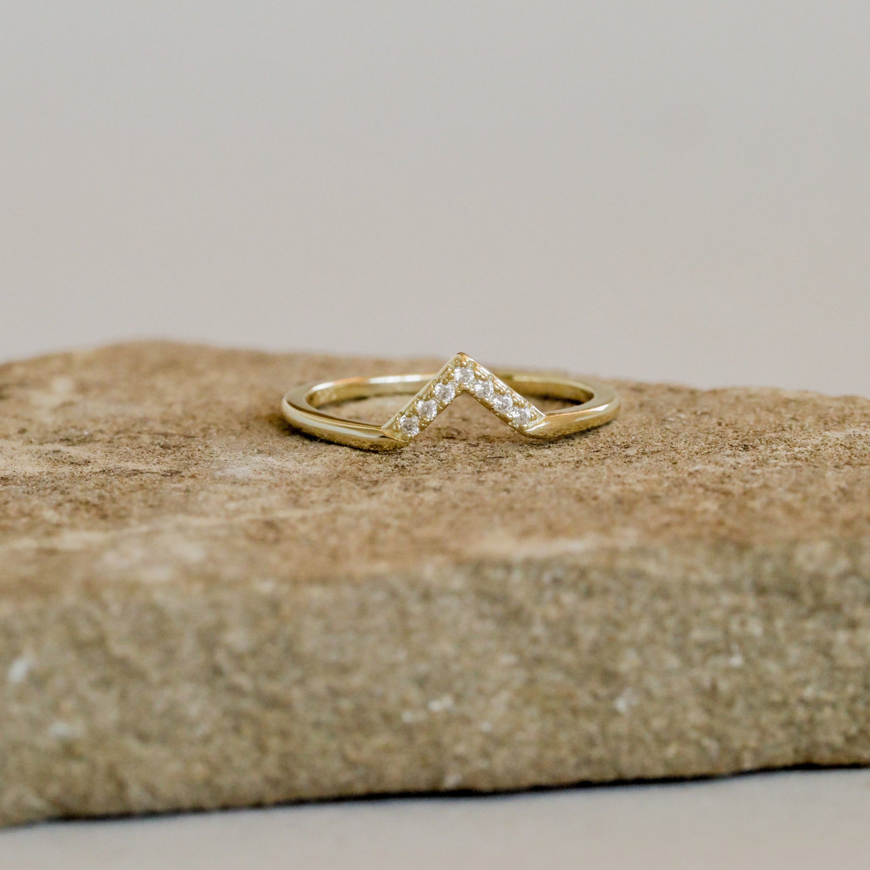 The Lambda Ring is a delicate gold-plated v-shaped ring with a chevron design and diamond accents, shown on a textured stone surface.