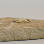 The Lambda Ring is a delicate gold-plated v-shaped ring with a chevron design and diamond accents, shown on a textured stone surface.