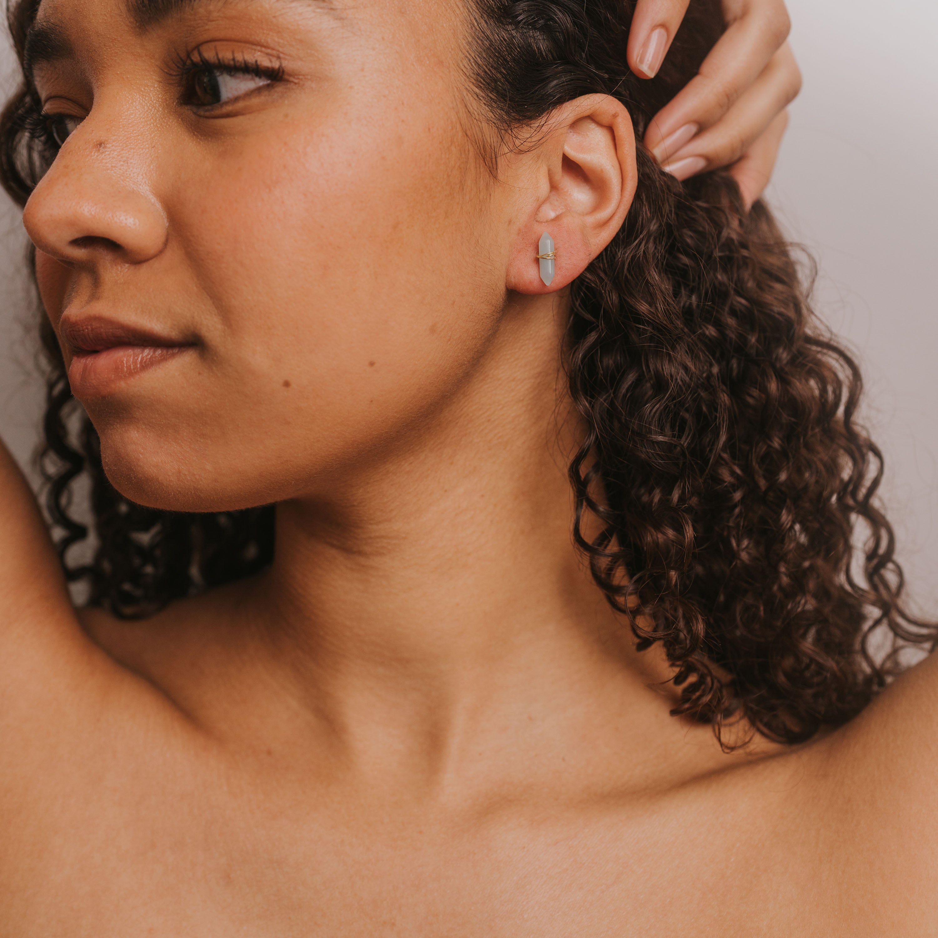 A woman with curly hair poses with one arm raised, wearing the Amazonite Mineral Point - Hope earring on her left ear. Her clear skin and neutral expression stand out against a soft, light background.