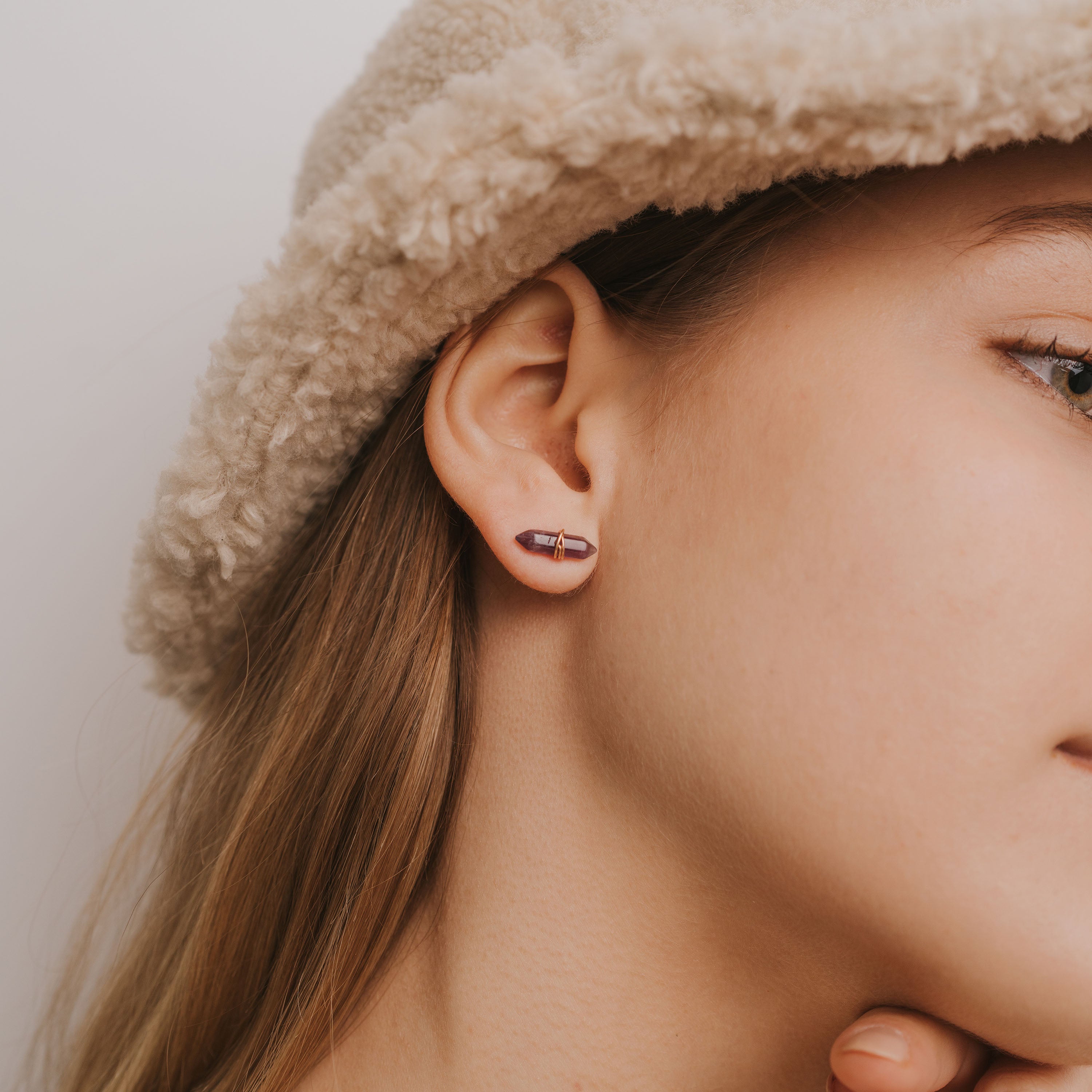 Close-up of a woman's ear wearing the Amethyst Mineral Point - Healing earring with sterling silver posts. She touches her chin and wears a beige textured hat, set against a plain, light-colored background.