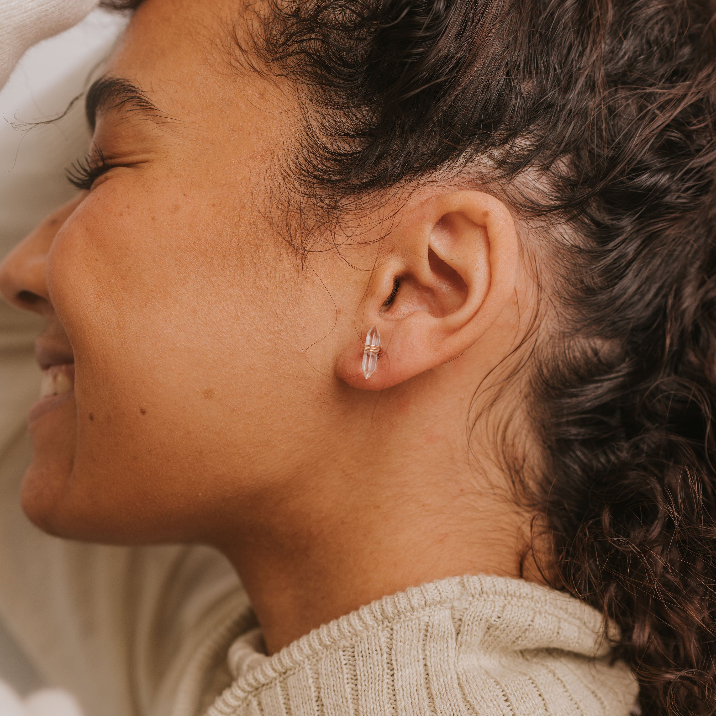 A close-up side view of a smiling person with curly hair wearing a Clear Quartz Mineral Point - Cleansing earring and a beige knit sweater, their eyes closed in peaceful contentment.