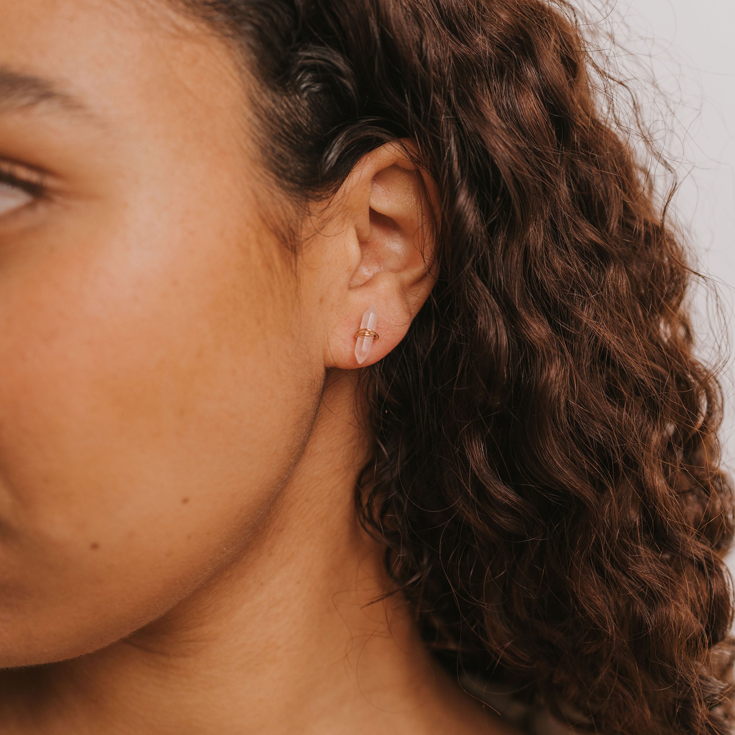 A close-up of a hand holding the Rose Quartz Mineral Point - Love crystal. The person has medium skin and curly brown hair. Soft lighting and a neutral background highlight the pink hues of the mineral point.